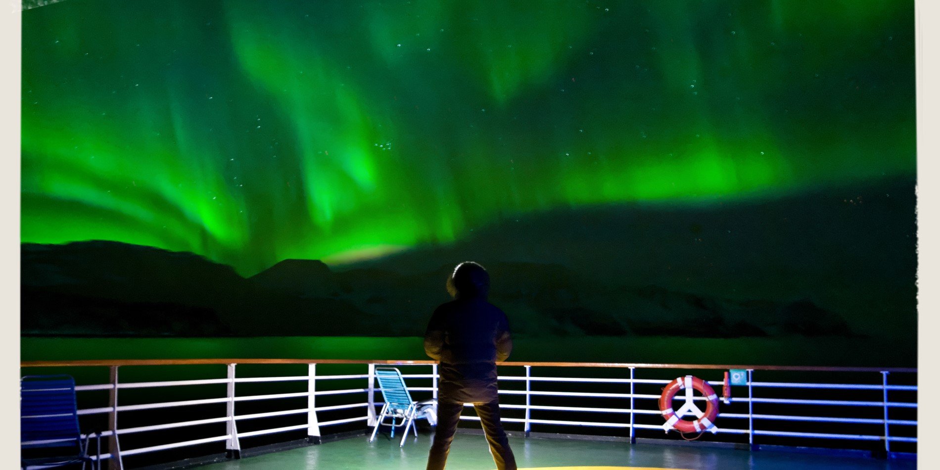 Man standing on deck of The Hurtigruten Norwegian Coastal Express. Above him is the sky lit up in green - the aurora is dancing