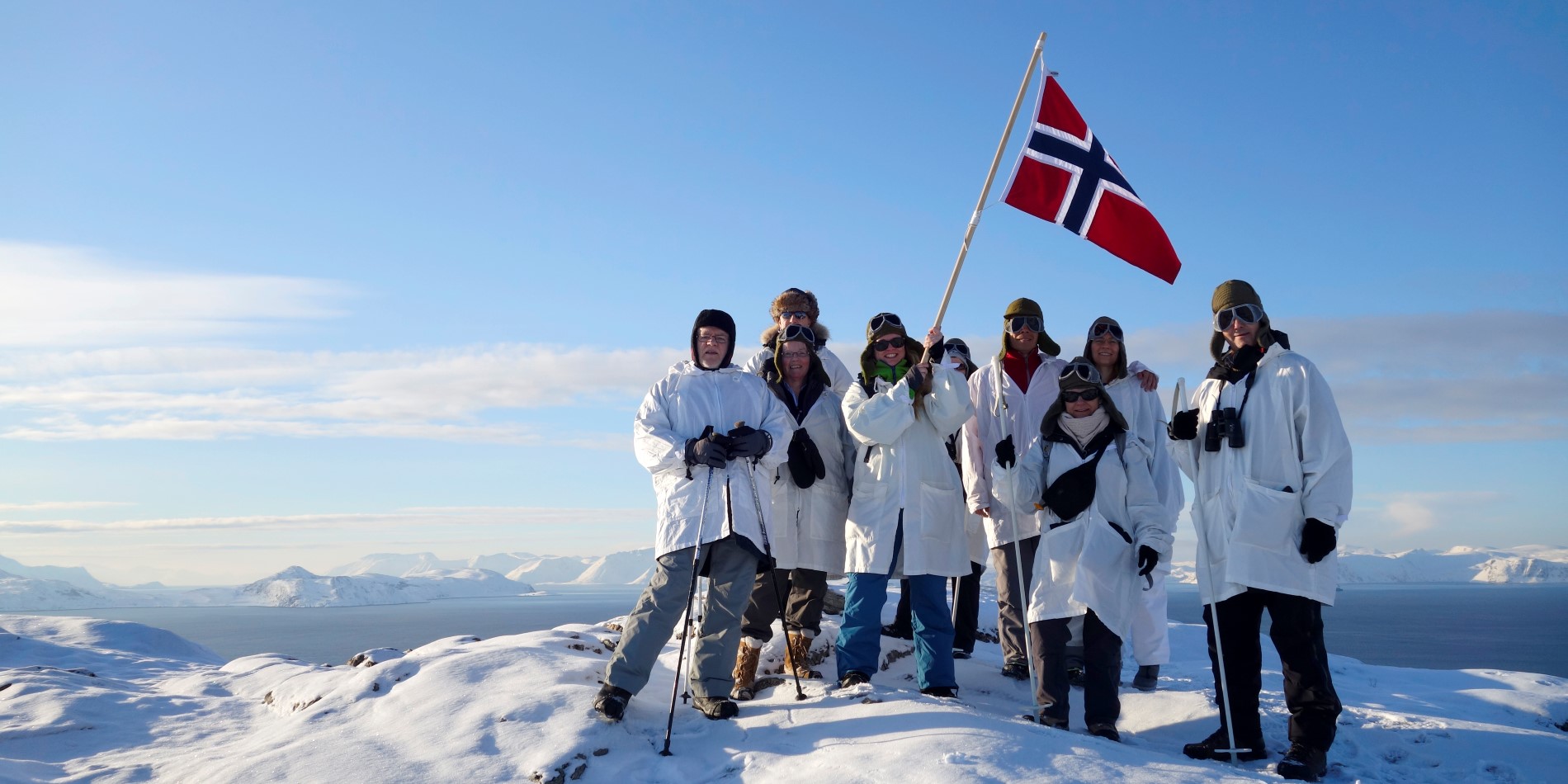 A group of 8 people in matching white jackets are waiving a Norwegian flag on top of a mountain top in Northern Norway
