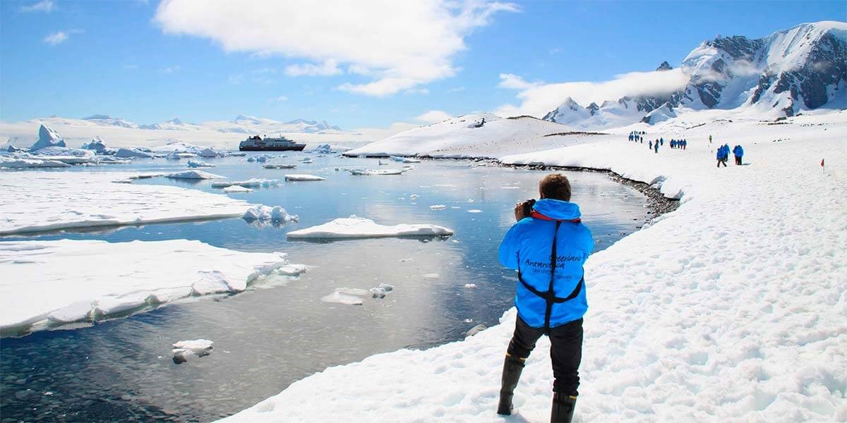 Man standing on the ice in Antarctica. Many persons are walking on the ice in front of him. Hurtigruten ship in the distance
