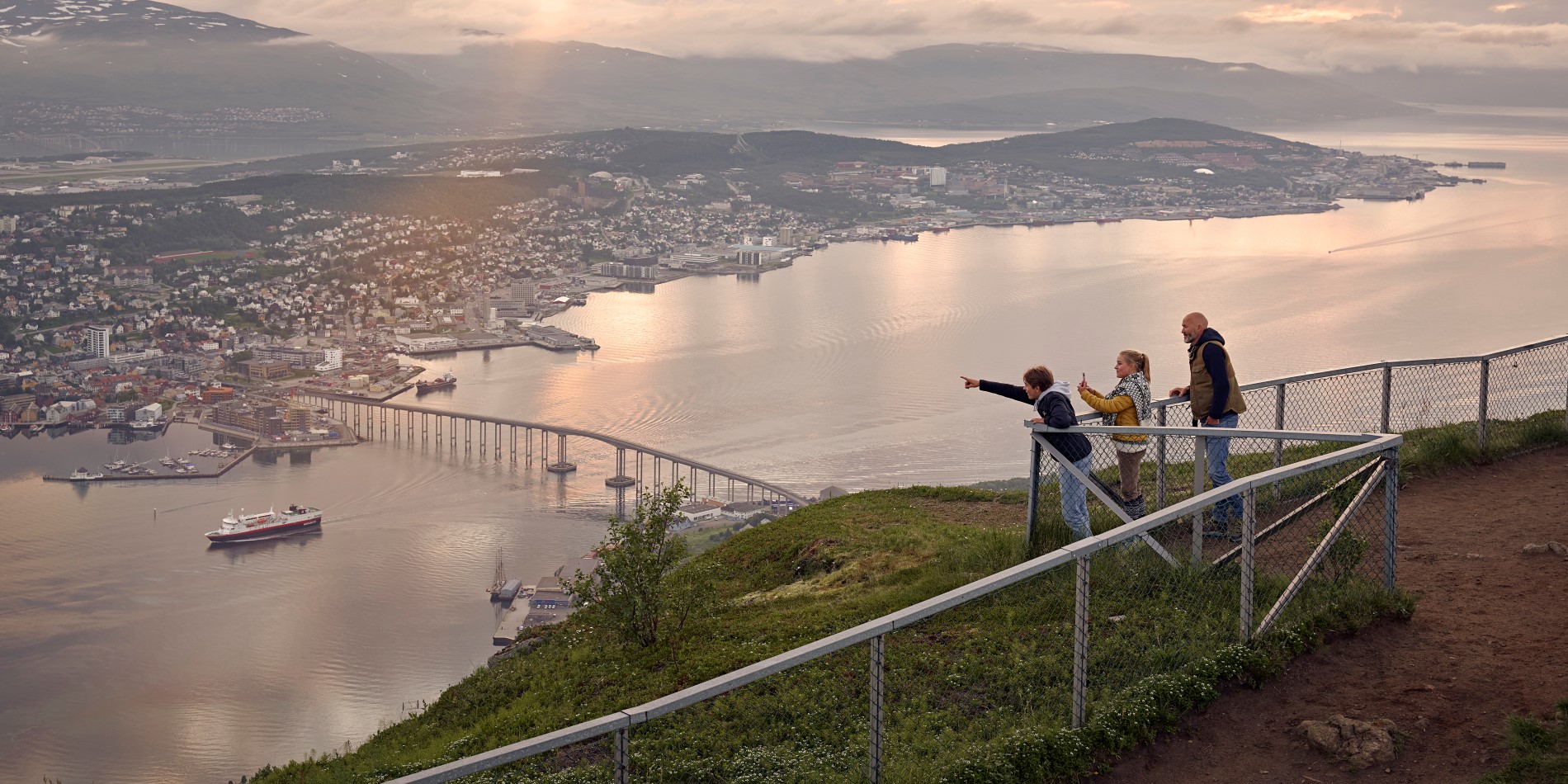 Three children standing behind the fence on top of the city mountain in Tromsø. They are overlooking Tromsø city.
