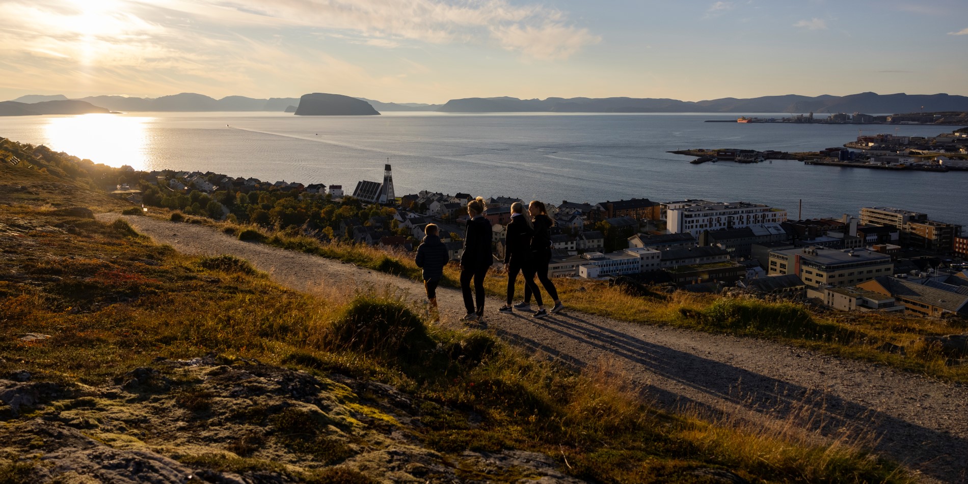 It's sunset and four people are walking in the hillside. Below they you can see the town of Hammerfest