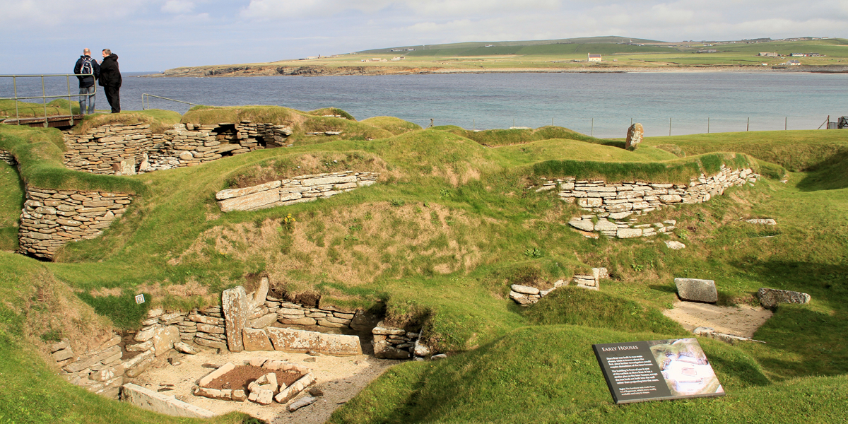 A group of people standing next to a body of water with Skara Brae in the background