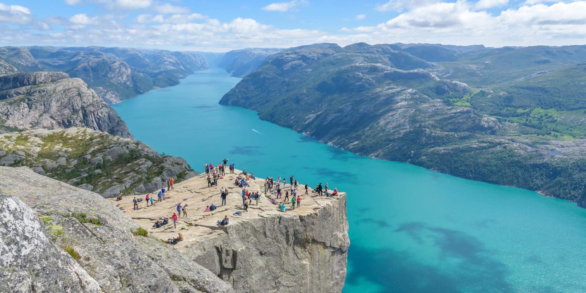 A group of people on a rock next to water with Preikestolen in the background