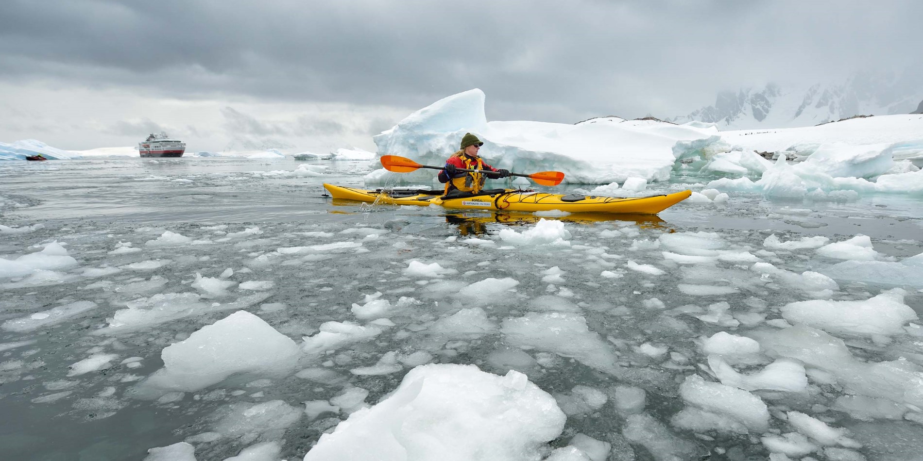 Woman kayaking among drift ice with explorer ship in background